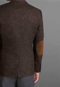 Giacca in tweed marrone con toppe in suede sui gomiti, vestibilità slim e dettagli ai polsi con bottoni. La vista posteriore mostra la spacco e la forma su misura.