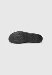 Semelle noire d'une chaussure avec une surface texturée présentant un motif en vagues et une marque. Le matériau semble en caoutchouc et durable.