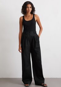 Zwarte geribbelde tanktop met brede, zwarte, hoge taille broeken. De broeken hebben een knoopsluiting en lichtjes textielachtige stof. Zwarte sandalen.