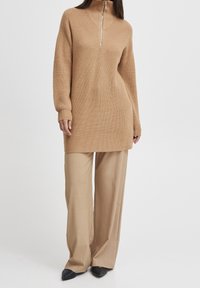 Maglione tunica lavorato a maglia di colore marrone chiaro con colletto alto e zip, caratterizzato da un motivo testurizzato. Abbinato a pantaloni beige a gamba larga e tacchi neri.