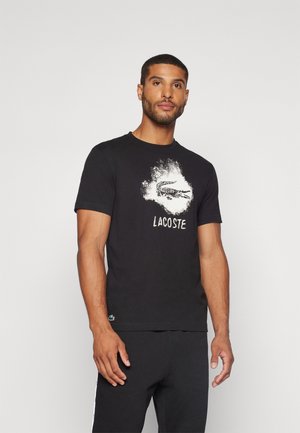 Svart bomullströja med vit grafisk print av en krokodil och texten "LACOSTE", med korta ärmar och rundad halsringning.