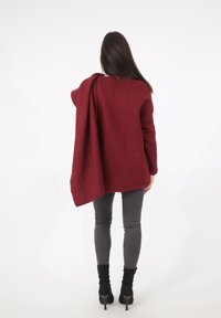 Maglione oversize bordeaux con texture appoggiato su una spalla, abbinato a leggings grigi aderenti e stivaletti neri con tacco alto.