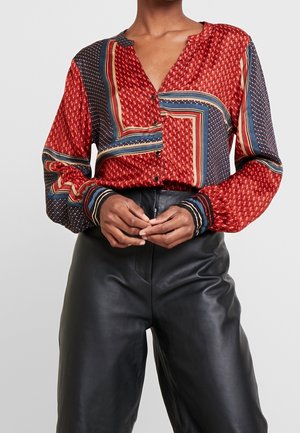 Femme portant un chemisier à motifs rouges, bleus et beiges, boutonné, à manches longues, associé à un pantalon en cuir noir taille haute.