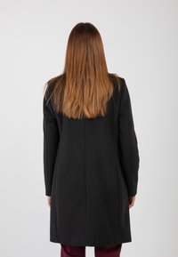 Persona con lunghi capelli castani lisci, che indossa un cappotto nero e pantaloni bordeaux, di spalle su uno sfondo grigio chiaro uniforme.