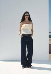 Top strapless bianco a balze, jeans blu scuro a gamba larga, occhiali da sole neri e sandali neri; si trova davanti a uno sfondo grigio chiaro.