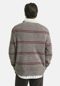 Pull en maille grise avec des rayures rouges, ourlet côtelé et tissu texturé. Porté sur une chemise blanche à col avec de fines rayures rouges.