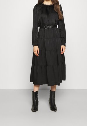 Kobieta ubrana w czarną, długorękawową sukienkę midi z dekoltem ozdobionym łańcuszkiem, paskiem w talii, warstwową spódnicą oraz czarnymi botkami do kostek.