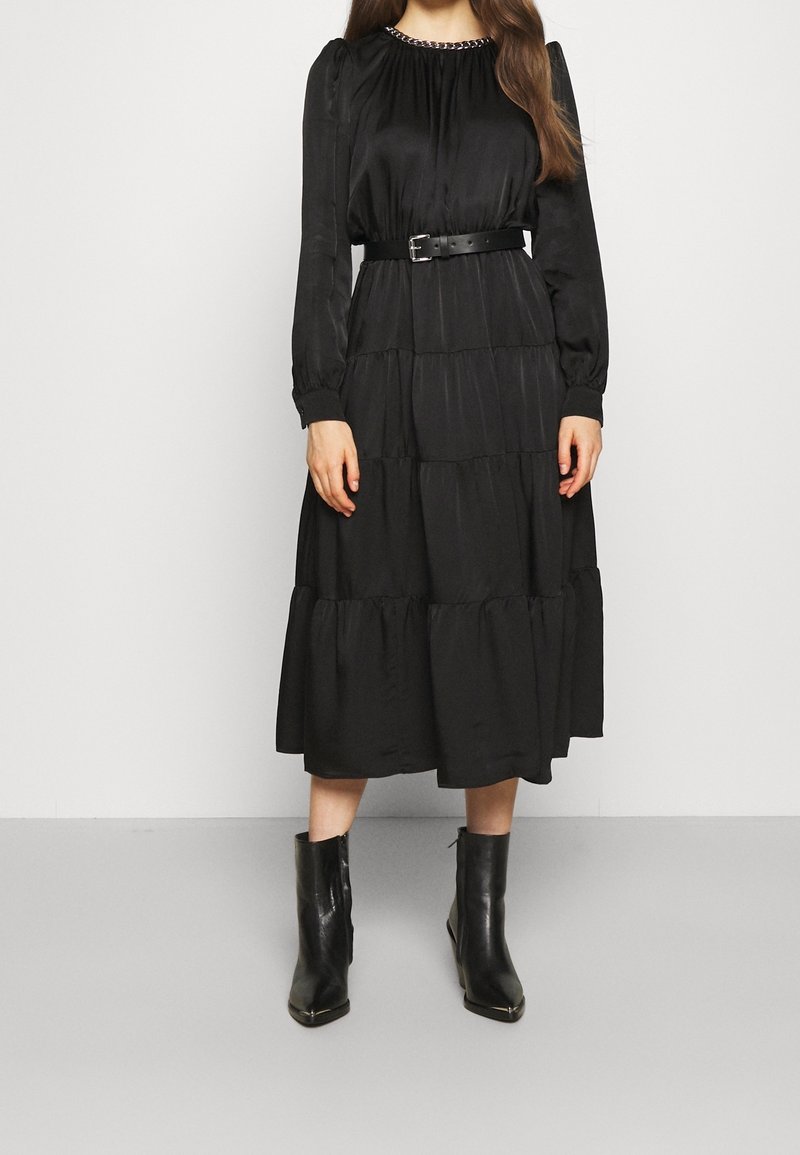 Femme portant une robe midi noire à manches longues avec un col en chaîne, une taille ceinturée, une jupe à volants et des bottines noires.