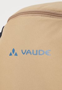 Beige stoffryggsekk med en teksturert overflate. Har en beige glidelås og en blå "VAUDE"-logo med en trekantet pynt.