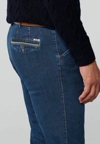 Jeans en denim bleu foncé avec une coupe ajustée, dotés d'une poche arrière avec fermeture éclair et d'un détail en bouton. Tissu texturé avec des coutures visibles.