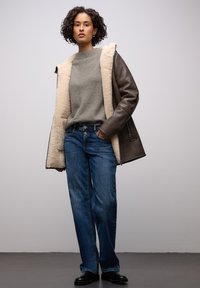 Ruskea nahkatakki lammasvuorella, harmaa teksturoitu neule ja siniset leveälahkeiset farkut. Mustat kengät, seisomassa neutraalia taustaa vasten.