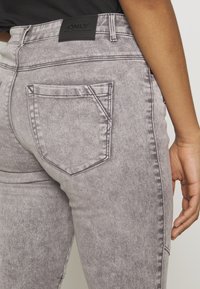Calças de ganga cinza com um acabamento texturizado, apresentando um único bolso traseiro e um patch de marca em pele preta na cintura.