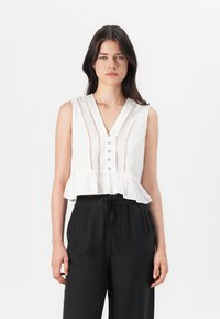 Hvid ærmeløs bluse med V-hals, frontlukning med knapper og flæsefortil; har lodret stribet tekstur og blonde detaljer. Båret med sorte bukser.
