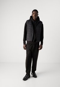 Black padded vest over a black hoodie, paired with loose black trousers and black shoes. The vest features zip closure and a structured design.