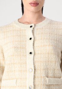 Béžový pletený cardigan s texturovaným vzorem, kulatým výstřihem, šesti knoflíky včetně černobílých akcentů a dvěma předními kapsami.