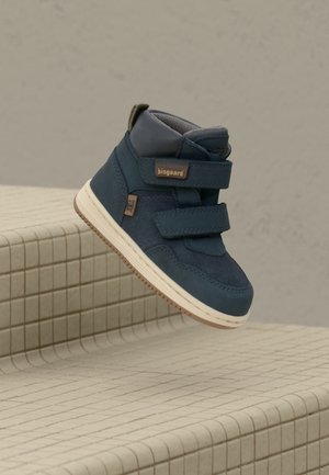Sneaker alta blu navy con cinturini in velcro, accenti contrastanti in beige, parte superiore testurizzata e suola in gomma bianca, posizionata su una superficie piastrellata.
