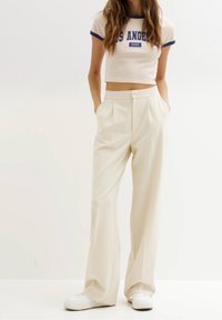 Pantalones de tiro alto en color beige claro, con piernas anchas, pliegues frontales y bolsillos laterales, combinados con una camiseta corta en color crema con ribete azul.