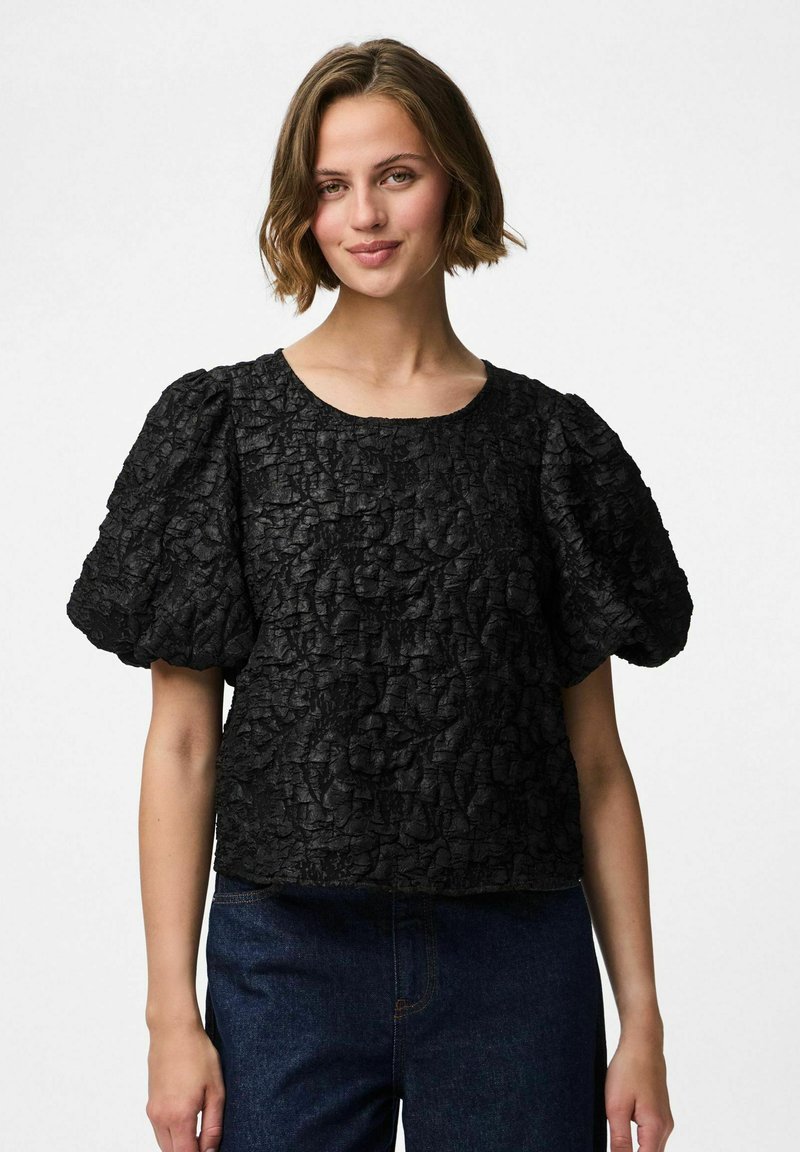 Svart texturerad topp med puffade korta ärmar och rund halsringning. Tyget har en krinklad design, kombinerat med mörka denimbyxor.