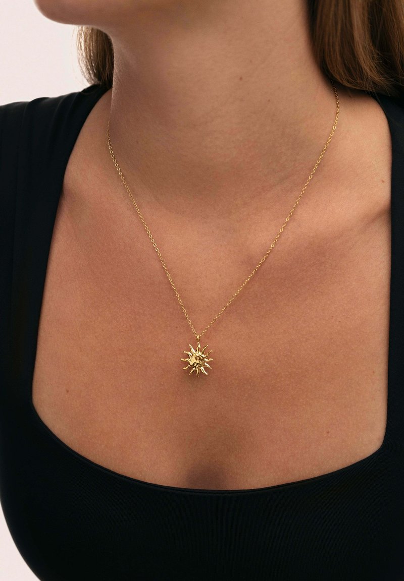 Collier en or avec un pendentif en forme de soleil aux rayons texturés, suspendu à une chaîne délicate contre une peau nue et un haut noir.