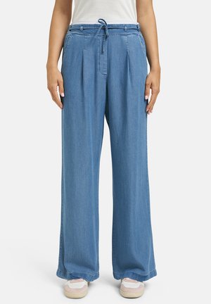 Woman wearing wide-leg light blue denim pants with front pleats, drawstring waist, white top, and white sneakers with pink accents.