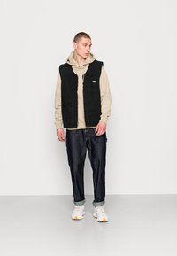 Black sleeveless vest with pockets layered over a beige hoodie, paired with dark denim pants and light sneakers. Simple, utilitarian design.