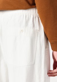 Hvite teksturerte bukser med elastisk livlinje, med en flat baklomme som er sikret med to knapper. Brukt med en brun langermet skjorte.