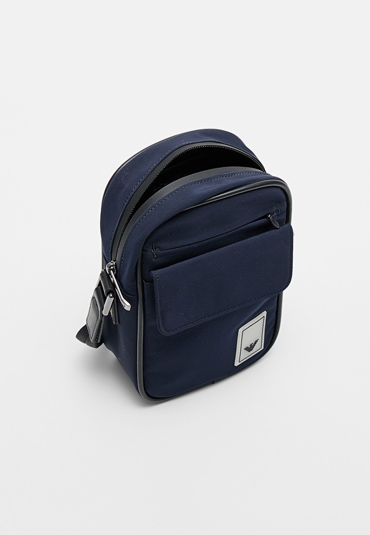 Bolso de nylon azul marino con textura suave, cierre doble de cremallera, bolsillo frontal con solapa y parche con logo. Forma compacta y rectangular.