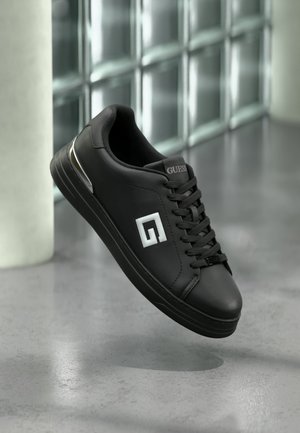 Zwarte leren sneaker met wit "G" logo en "Guess" op de tong, hangend boven een reflecterende grijze vloer bij een glasblokkenmuur.