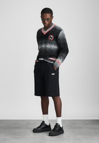 Čierny a šedý gradientový sveter s pleteným vzorom a červenými akcentmi, výstrih do V, s logom, skombinovaný s čiernymi šortkami na šnúrku a čiernymi teniskami.