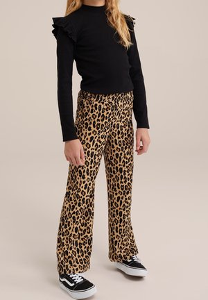 Kind trägt schwarzen Langarmtop mit Rüschenschultern, Leopardenmuster-Hose und schwarz-weißen Turnschuhen und steht vor beigem Hintergrund.