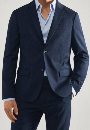 Uomo che indossa un abito blu scuro con una camicia azzurra, una mano in tasca e l'altra rilassata di fianco.