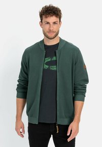 Grüne gerippte Strickjacke mit Reißverschluss, die über eine gestrickte Textur, gerippte Bündchen und einen braunen Lederakzent am Ärmel verfügt. Getragen über einem schwarzen T-Shirt.