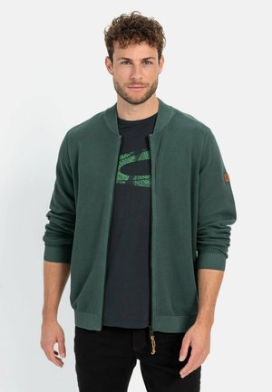 Grüne gerippte Strickjacke mit Reißverschluss, die über eine gestrickte Textur, gerippte Bündchen und einen braunen Lederakzent am Ärmel verfügt. Getragen über einem schwarzen T-Shirt.