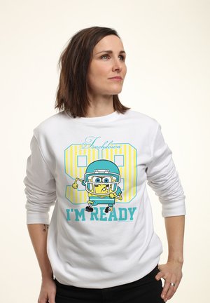 Kobieta o brązowych włosach nosi białą bluzę z nadrukiem kreskówkowego zawodnika futbolu amerykańskiego, numer 89, oraz napisem „Touchdown I'm Ready”, patrząc na bok.