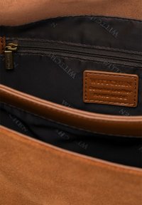 Wnętrze brązowej skórzanej torby z kieszenią na zamek i metką z logo. Podszewka jest z materiału z powtarzającymi się wzorami brandingowymi. Gładka tekstura.