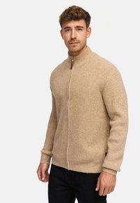 Beżowy sweter zapinany na zamek, wykonany z prążkowanej dzianiny, z wysokim kołnierzem, brązowym skórzanym uchwytem zamka i długimi rękawami. Miękka tekstura i dopasowany krój.
