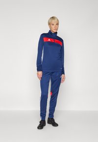 Navy athletic tracksuit featuring a red accent stripe, zip-up collar, and white side detailing. Made from smooth synthetic fabric.