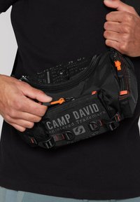 Black fabric waist bag with multiple zippered pockets, orange zipper pulls, and printed logo. Features adjustable straps and clip hardware.
