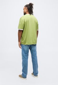 Άνδρας με dreadlocks που φοράει πράσινο μπλουζάκι με κοντά μανίκια, μπλε τζιν και μπεζ αθλητικά παπούτσια, στέκεται με την πλάτη προς το μέρος μας μπροστά σε μια απλή φόντο.