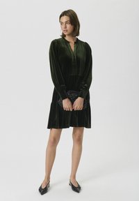 Grünes Samtkleid mit einem Kragen, langen Ärmeln und einer gerafften Taille. Das Model hält eine schwarze Clutch und trägt schwarze spitze Flats.