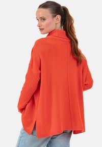 Pull tricoté orange surdimensionné avec un col roulé, des manches côtelées et des fentes latérales, présentant une coupe décontractée et une texture douce.