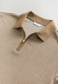 Chaleco con cuello de cremallera en color marrón claro con un patrón de cuadros texturizado. Presenta un cuello acanalado y un lazo de tela para facilitar su uso. Acabado interior suave.