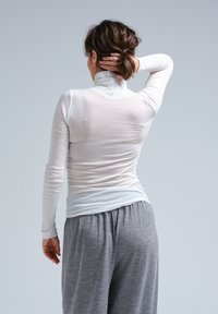 Hvid langærmet top med høj krave, båret over lysegrå, løstsiddende bukser. Stoffet ser strækbart og tyndt ud.