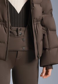 Smeđa pufer jakna na patentni zatvarač s velikim prešivanim dijelovima, nošena preko tamnog rebrastog topa i usklađenih hlača s visokim strukom i drukerima.