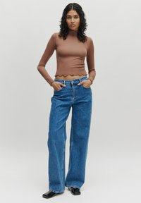 Bruine lange mouwen crop top met een gegolfde zoom, gecombineerd met hooggeglyde, wijde spijkerbroek in het blauw. Model staat met handen in de zakken.