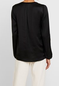 Blusa nera di seta con maniche lunghe, collo rotondo e una piega centrale sul retro. Tessuto morbido e vestibilità rilassata.