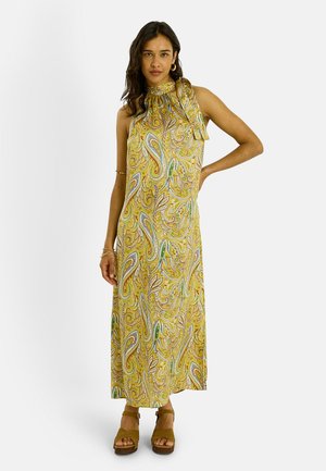 Mujer con un vestido maxi amarillo sin mangas con estampados coloridos de paisley, de pie con una mano en la cadera, llevando sandalias marrones con plataforma.