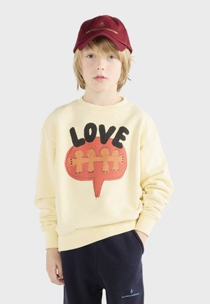 Crème sweatshirt met zwart en oranje "LOVE" grafisch ontwerp, gecombineerd met donkere sweatpants en een bordeauxrode pet met gouden tekst.