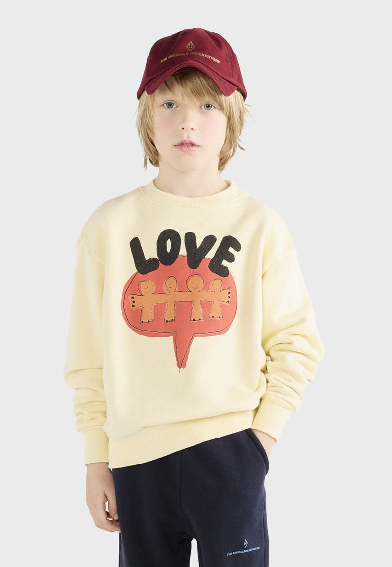 Krem sweatshirt s grafikom "LOVE" u crnoj i narančastoj boji, uparen s tamnim duksericama i bordo kapom s zlatnim natpisom.