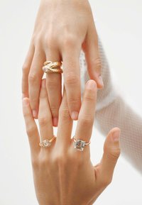 Tres anillos exhibidos en los dedos: dos bandas de oro con patrones trenzados y un anillo con un stone rectangular. Brillo notable y texturas suaves.